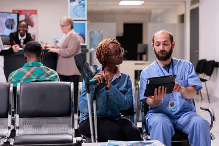 Medical nurse showing diagnosis to female patient with crutches, explaining disease and treatment on digital tablet. Specialist talking to woman with impairment in hospital reception.の写真素材