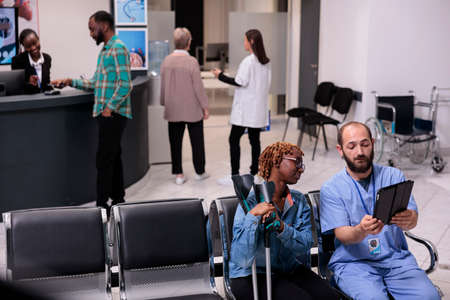 African american patient and nurse looking at tablet to find dsease diagnosis on digital device, suffering from physical impairment in waiting area. Woman and specialist in hospital lobby.の写真素材