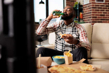 Young adult drinking alcoholic beer from bottle and eating takeaway meal from fast food delivery...