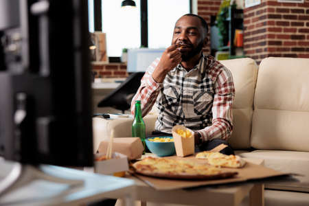 African american man eating chips while he watches film on tv, having fun with takeaway delivery and beer bottle. Watching movie on television and enjoying takeout fast food meal at home.の写真素材