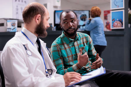 General practitioner talking to male patient in waiting room, using medical checkup report to give disease diagnosis and treatment. Doctor and man doing healthcare consultation.の写真素材