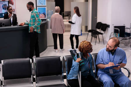 Health specialist consulting woman with crutches in hospital reception lobby, talking about diagnosis and medicine in waiting room area. Diverse people discussing at checkup appointment.の写真素材