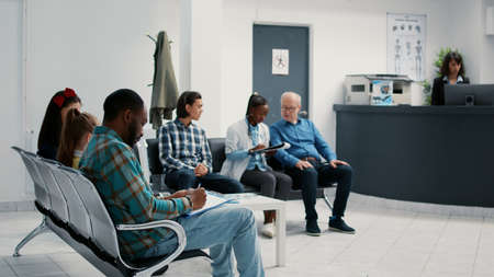 Diverse group of patients sitting in waiting room at hospital, reception desk for checkup appointment and consultation. Waiting area lobby with people at medical examination facility.の写真素材