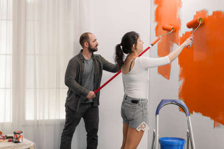 Young couple using orange color to paint wall with roller and paintbrush during home renovation....