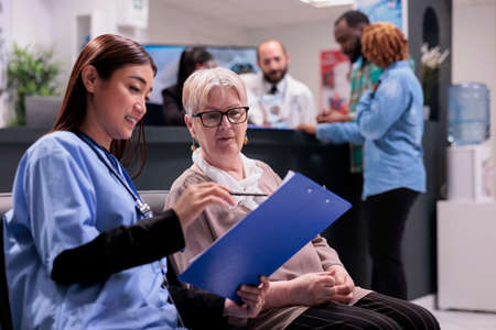 Asian nurse explaining diagnosis to elderly patient, sitting in waiting room lobby and talking about...