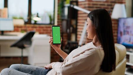 Young adult person sitting on couch with mobile cellphone having mockup chroma key background. Asian woman having smartphone with green screen display while sitting at home on sofa. Tripod shotの写真素材