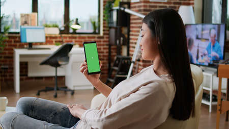 Happy smiling heartily woman having smartphone with green screen display while sitting at home on...