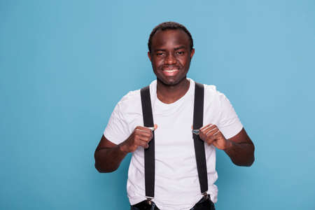 Happy young man pulling suspenders on blue background. Confident handsome african person wearing white tshirt and black suspenders while smiling heartily at camera. Studio shotの写真素材