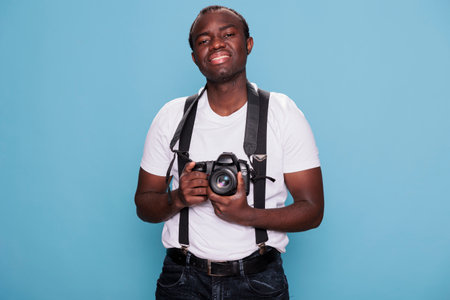 Joyful handsome looking african ethnicity guy with photo device posing with confidence. Confident professional photographer having modern camera while smiling heartily and standing on blue background.の写真素材