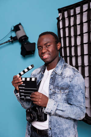 Smiling photographer with clapper on blue background while having softboxes and spotlights. Young...