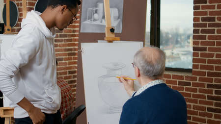 Young artist explaining drawing technique to elder person, using pencil to develop artistic skills on canvas and easel. Senior student learning to draw vase model in art class with teacher.の写真素材
