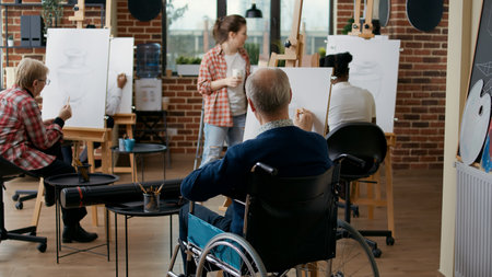 Old man with disability using pencil to draw masterpiece in art class with people. Elder student in wheelchair drawing vase model on canvas, working with artistic tools for personal development.の写真素材