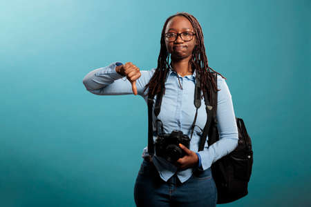 Disapproving photographer giving thumbs down gesture at camera while standing on blue background. Young adult photography entusiast with DSLR device showing dislike hand symbol.の写真素材