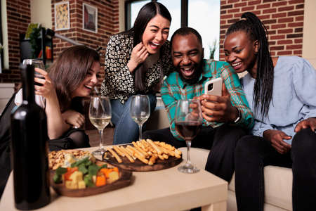Diverse group of friends at wine party celebrating birthday while watching funny clips on smartphone. Happy young people having a glass of wine and eating snacks while enjoying time together.の写真素材