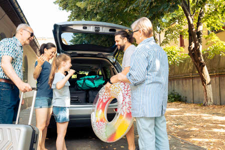 Happy people travelling at seaside with vehicle, leaving on vacation trip with parents, grandparents and small girl. Loading luggage and suitcase in automobile trunk, summer adventure.の写真素材