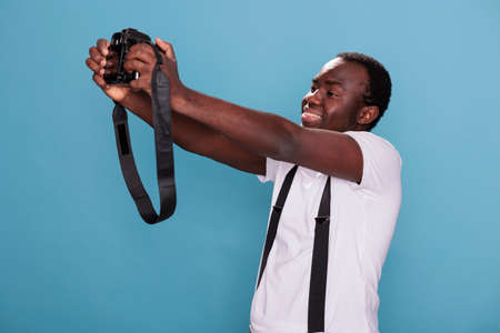 Happy professional photographer taking selfie photo with modern camera while standing on blue background. Smiling heartily photography enthusiast taking picture of himself. Studio shot.の写真素材