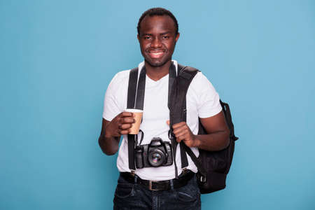 Tourist with professional camera and travel backpack ready for holiday journey on citybreak. African american photographer with DSLR photo device and travel roadtrip bag standing on blue background.の写真素材