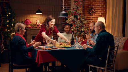 Lovely housewife bringing main course to Christmas dinner table. Impatient and happy festive family members gathered at home to celebrate winter holiday with traditional home cooked food.の写真素材
