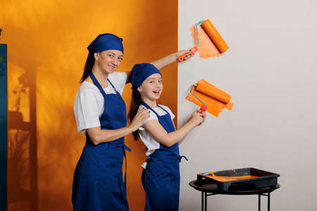 Portrait of family using orange paint to change walls color in apartment room, painting with roller brush to renovate house. Woman with small kid doing diy home redecoration together.の写真素材