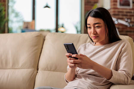 Casual authentic looking happy asian woman using smartphone at home, web internet technology smartphone user mobile message social media communication digital deviceの写真素材