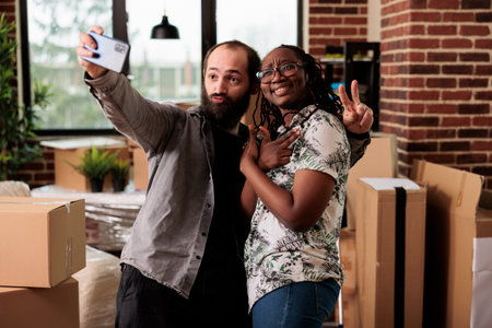 Diverse married couple having fun with pictures on smartphone, recording video of moving in new...