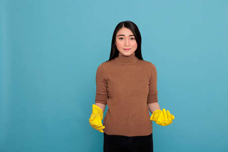 Happy smiling asian housewife from chores wearing yellow gloves standing against a blue background, Cleaning home concept, Cheerful satisfied and glad with positive state of mindの写真素材