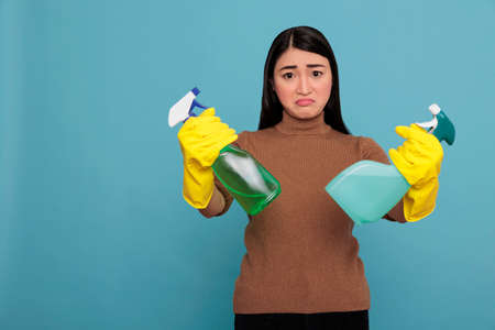 Sadly angry young asian woman holding two detergent spray in yellow gloves isolated on a blue background, Cleaning home concept, Unhappy depressed and exhausted negative housekeeperの写真素材