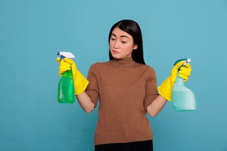 Portrait of pretty sad asian woman holding two detergent sprayers in yellow gloves isolated on a blue background, Cleaning home concept, Unhappy depressed and negative housewifeの写真素材