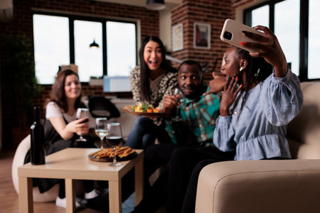 Happy smiling adults at home enjoying time together while drinking wine at home. Diverse cheerful young people in living room celebrating birthday party while taking group pictures.の写真素材