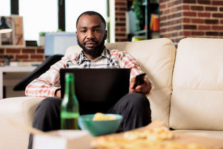 Casual relaxed person working on laptop and switching tv channels with remote control, browsing internet and finding film to watch. Eating takeout delivery fast food meal at home.の写真素材