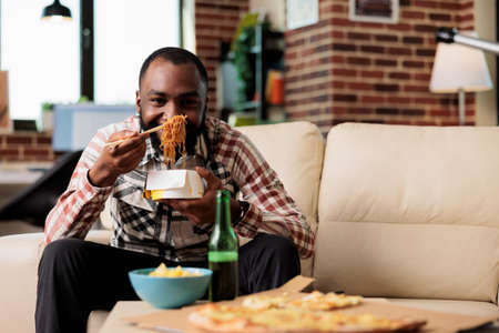 African american man using chopsticks to eat takeout noodles food at home, watching movie on tv channel. Eating takeaway delivery meal from box and enjoying film on television.の写真素材