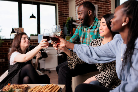 Happy diverse friends clinking wineglasses at birthday party in living room. Joyful group of multiethnic people toasting at wine party while celebrating friendship event.の写真素材