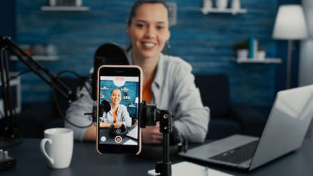 Attractive social media influencer sitting at home studio desk while starting streaming from living room with smartphone. Young content creator recording daily vlog with touchscreen modern device.の写真素材