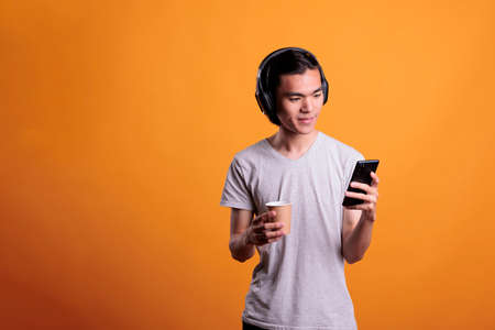 Man in wireless headphones listening to podcast on mobile phone, holding coffee to go. Smiling asian teenager, using social media on smartphone, enjoying music in earphonesの写真素材