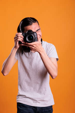 Photographer taking photos on professional digital camera, looking in viewfinder. Young asian man using photographing equipment, front view medium shot, creative hobby, photoshootの写真素材