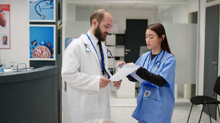 General practitioner and asian nurse talking about healthcare service, looking at medical report papers in hospital reception lobby. Diverse clinical staff discussing about treatment.の写真素材