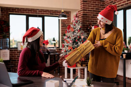 Asian woman receiving christmas present from coworker to celebrate xmas holiday in office with festive decorations and seasonal winter ornaments. Giving gifts to colleagues in business space.の写真素材