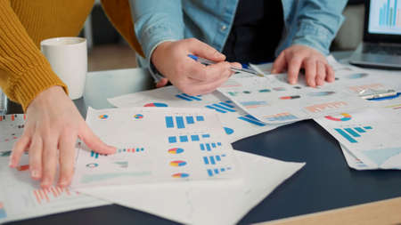 Selective focus on colleagues comparing marketing statistics and working on business strategy at desk with laptop. Detail view of coworkers hands going through papers with sales charts.の写真素材