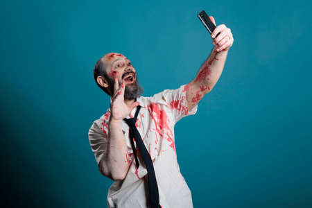 Apocalyptic corpse taking photo on smartphone, fooling around on camera and acting eerie. Spooky zombie with ugly wounds and bloody scars taking picture on mobile phone, brain eating.の写真素材