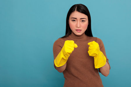 Sad angry asian young housewife in rubber gloves standing in a fight pose isolated on a blue background, Ready for his duty, Cleaning home concept, Female in aggressive face expressionの写真素材
