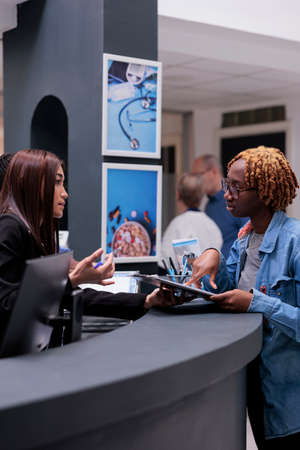 Female receptionist helping woman at reception desk, signing medical report papers to receive insurance support before checkup appointment. Patient filling in registration form to do examination.の写真素材