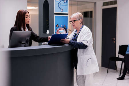 Senior doctor working with asian receptionist to plan checkup report papers at hospital reception desk. Medic taking to clinic worker at counter about medical forms and healthcare insurance support.の写真素材