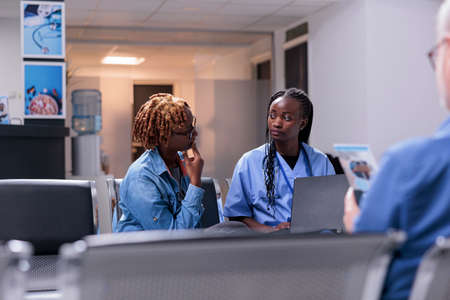 African american people doing checkup visit with disease diagnosis on laptop, sitting in waiting area lobby. Nurse and patient talking about treatment and recovery, healthcare support at clinic.の写真素材