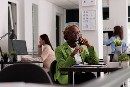 Employee talking on smartphone in coworking open space, having phone call with business partners. Young african american woman sitting at workplace desk, busy office workersの写真素材