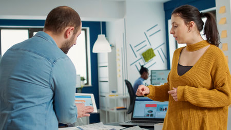 Man in startup office zoomin in digital tablet using touchscreen presenting sales charts to woman...
