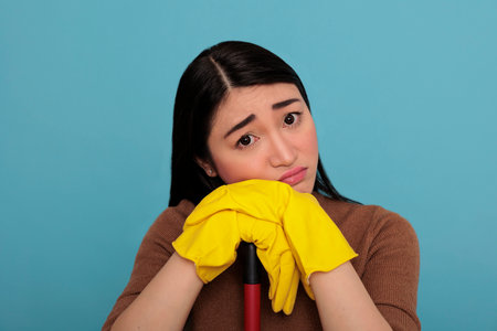 Overworked negative asian housemaid holding on brush stick yellow gloves holding on brush stick, Cleaning home concept ,Housekeeper feeling depressed exhausted taking some restの写真素材
