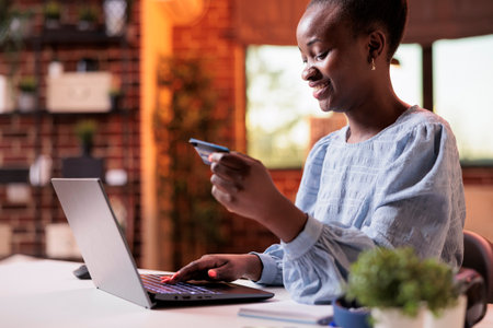 Smiling woman making purchase in internet shop using laptop. Young african american girl holding debit card and searching goods in online store, electronic shopping conceptの写真素材