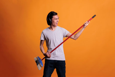 Young asian man in wireless, headphones playing rock song on mop as virtual guitar. Musician...