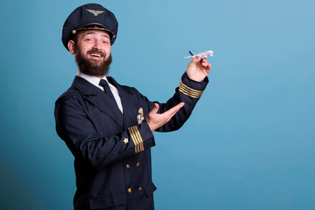 Smiling pilot with uniform playing with airplane model side view, aviation academy aviator holding commercial plane toy. Funny aircraft capitan, studio medium shot on blue backgroundの写真素材