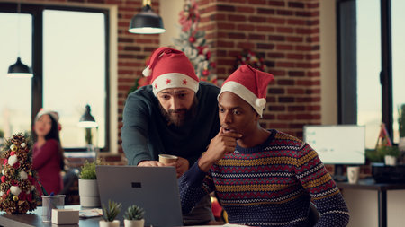 Multiethnic team of people analyzing report on computer during christmas eve festivity, wearing santa hats. Diverse men working together in office with xmas tree and seasonal ornaments.の写真素材